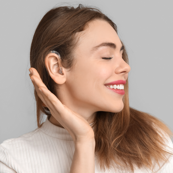 Uśmiechnięta kobieta z zamkniętymi oczami, ma odsłonięte prawe ucho, na którym założony jest aparat słuchowy.