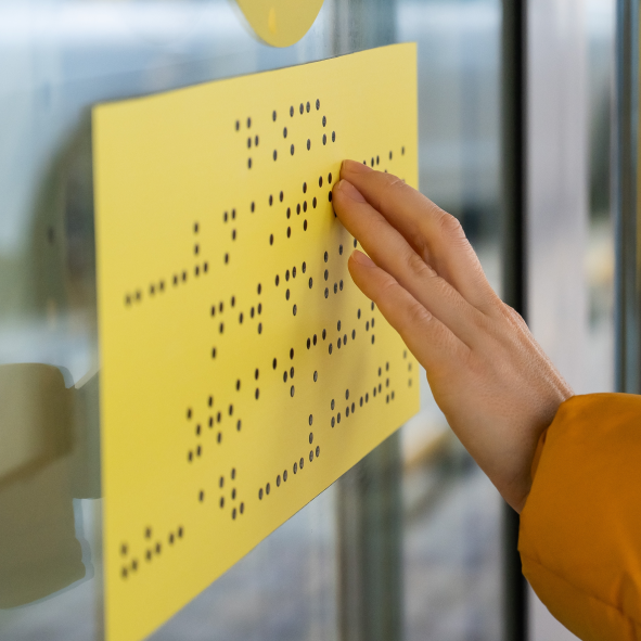 Na szybie wisi żółta kartka z alfabetem Braille’a, którą dotyka dłoń.