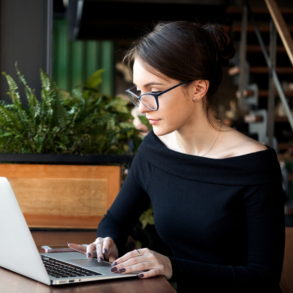 Kobieta w okularach z ciemnymi włosami spiętymi w kok siedzi i pracuje na laptopie.