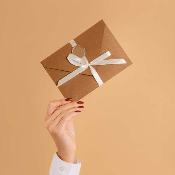damska ręka trzyma w ręku beżową kopertę przewiązaną wstążką białą